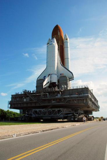 A space shuttle attached to a rocket is on a massive mobile crawler on a roadway.