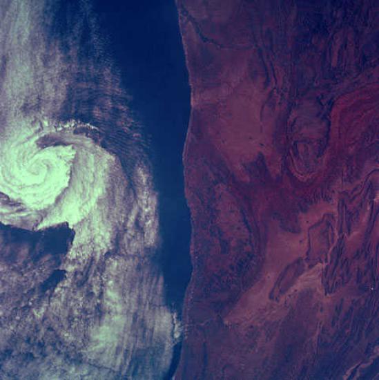 A satellite image. The right half shows the coast of a land mass and the left half shows a swirling storm over the ocean.