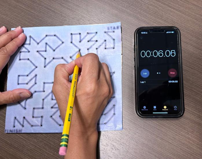 On the left side of the frame, a hand holding a pencil is completing a maze. On the right side of the frame, an iPhone is open to a stopwatch app.