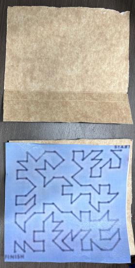 Two pieces of square paper on a table. One is a print out of a maze used by Skylab astronauts. One is blank.