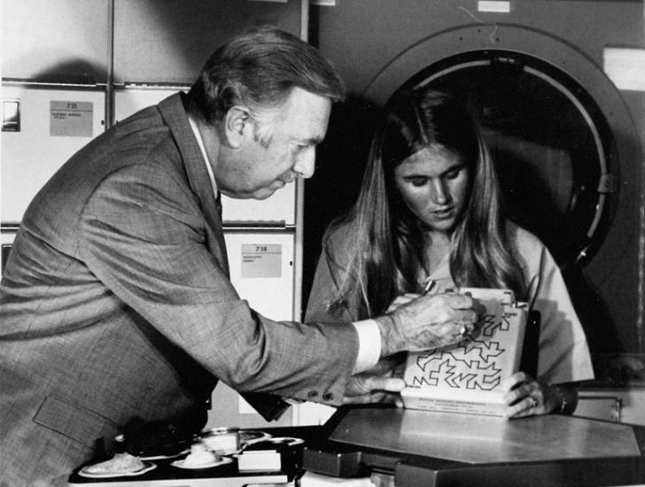 A black and white photograph of a young woman showing a maze to an older man in a suit.