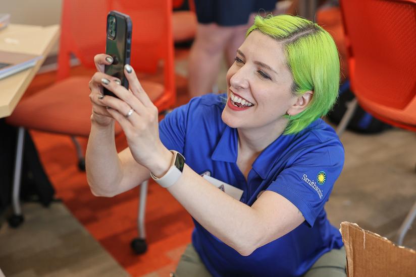 An educator in a blue polo smiles while taking a picture on her smart phone. 