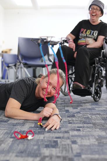 In the foreground, a small hovercraft or drone floats with pipe cleaner attached. Behind it, an educator lies on the floor smiling. Behind them, another educator, seated in a wheelchair, laughs. 