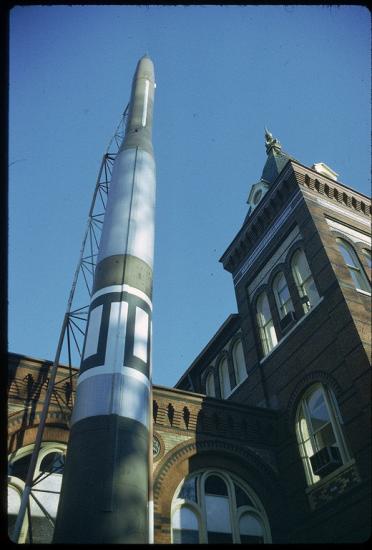 The top three-quarters of green and white missile seen from the ground looking up. The rocket sits vertically in front of an ornate building.