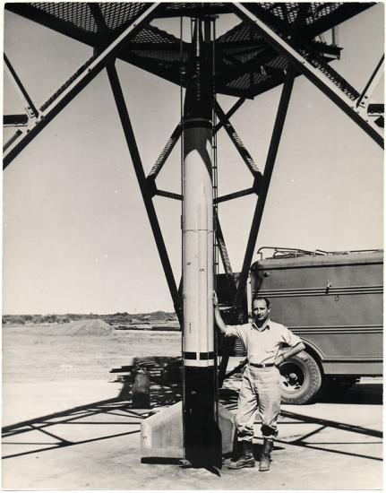 A man leans on a rocket that is about two times as tall as him. The rocket is fixed to a launch tower. Part of a truck can be seen in the background.