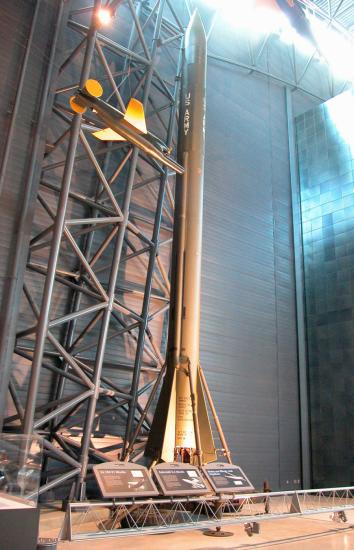 A large olive green rocket on display in a hangar with signage in front of it.