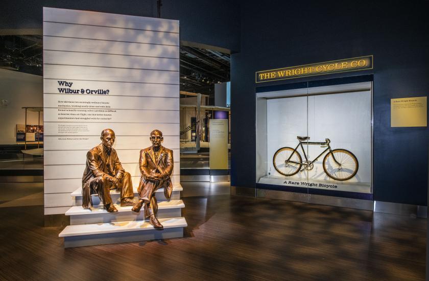 A photo looking into the entrance of a museum exhibit. A bicycle can be seen on display behind plexiglass and a statue of two men sitting adorn the entryway panel which says "who were wilbur & orville."