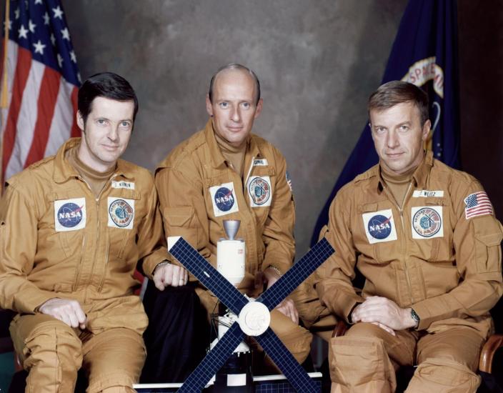 Three people in orange jumpsuits sit posed in front of two flags with a small model of Skylab in front of them.