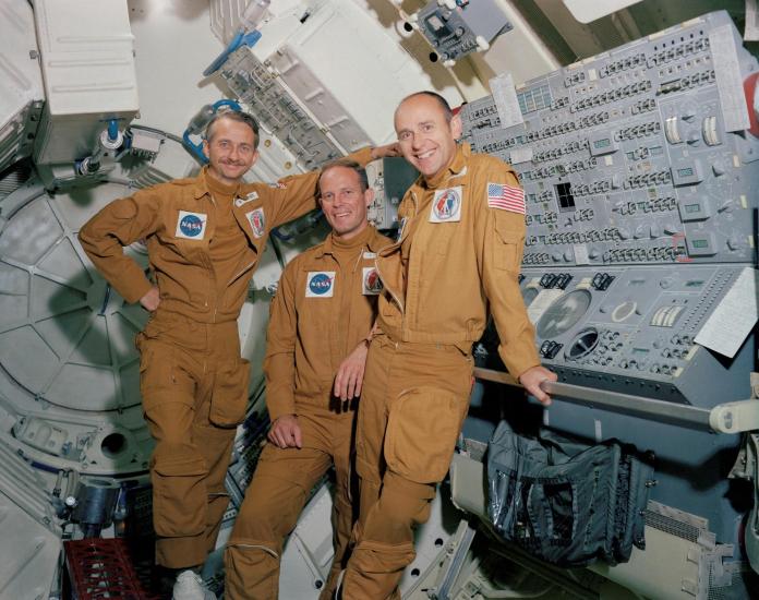 Three men stand facing the camera. The two on the righthand side lean against a control board inside a training module at NASA.