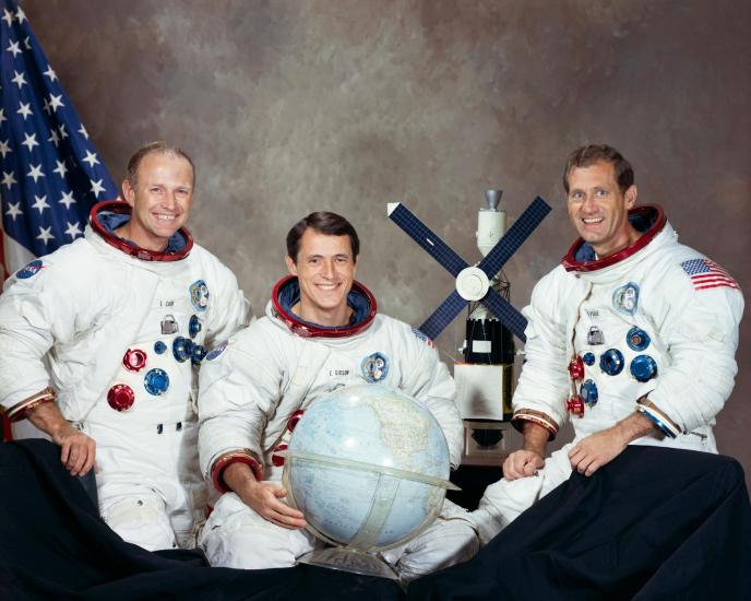 Three people seated against a neutral background with an American flag to the left. All three people are wearing spacesuits. The person in the middle holds a globe. A model of Skylab is situation between the person in the middle and the person on the right.