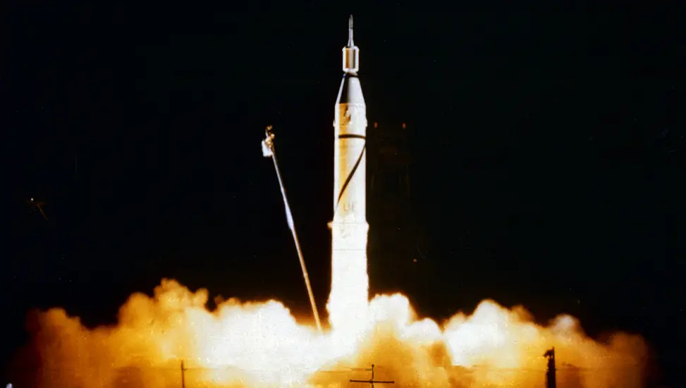 A large rocket taking off of a landing pad with a satellite attached to the top of it. Smoke and exhaust bellow out from the bottom of the rocket.