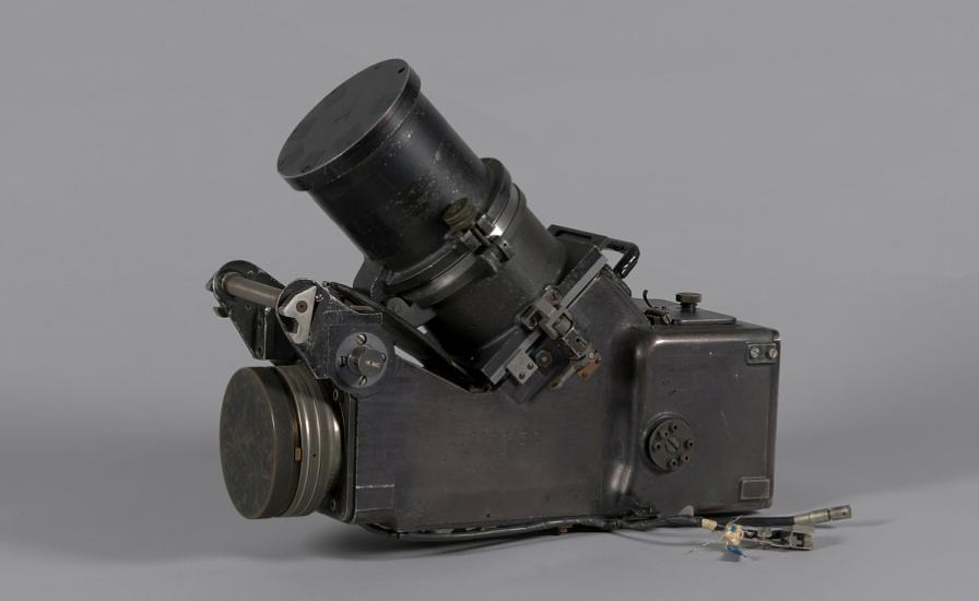 A black object similar in shape to a camcorder. It has a rectangular body with a lens on one end. Its also has another lens sticking out of the top.