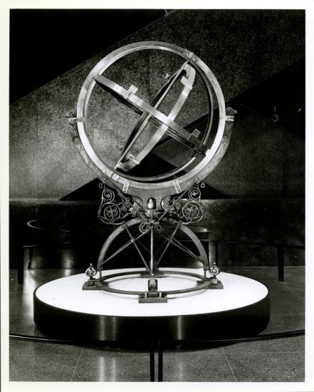 A black and white photo of a spherical sculpture with three sets of metal rotating rings. The center ring is the smallest and each ring after encompasses the one before it.