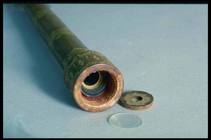 A photo of the end of a simple wooden telescope embellished with golden filigree painted on its surface. The lens and end cap are laid on the table next to it.