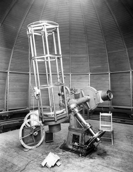 A black and white photograph of a large telescope inside of an observatory. A chair is situated next to it, the juxtaposition demonstrating that the chair is roughly 1/3 the size of the chair.