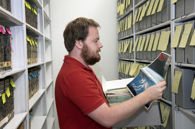 A man opens an archival box and holds a news magazine. On the magazine is a picture of Earth from space. Behind him are shelves of archival boxes. 