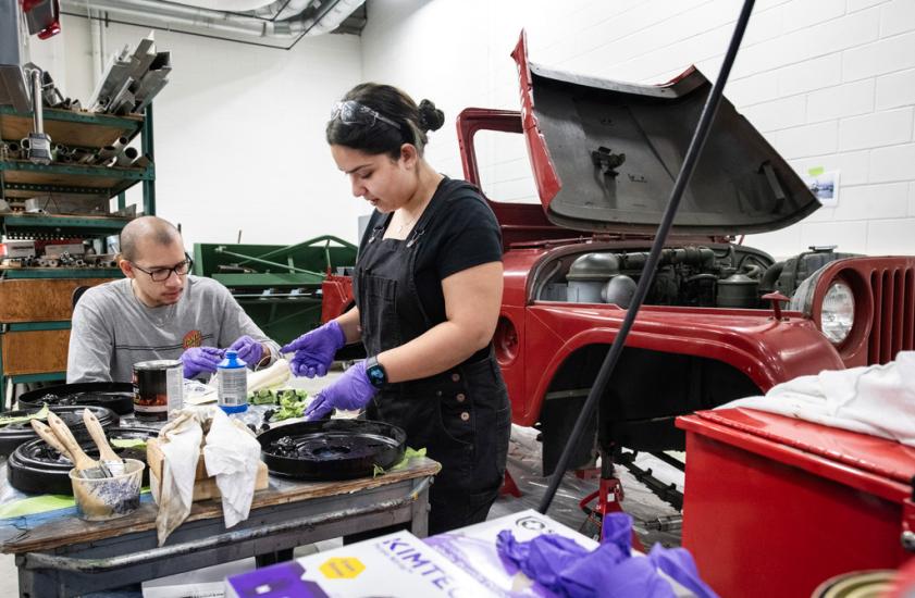 Two interns, a young man and young woman, work at a table wearing gloves and eye protection. Behind them is a jeep with the hood open. 