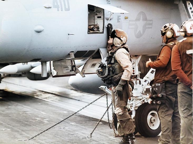 A young woman wearing a U.S. Navy flightsuit, helmet, goggles, and gloves peers into an open compartment on the side of a gray military jet with U.S. Navy markings.