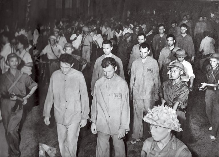 A black-and-white image of multiple American men walking down a street while wearing loose-fitting prison uniforms. The men are looking down as North Vietnamese soldiers and civilians press in closely.