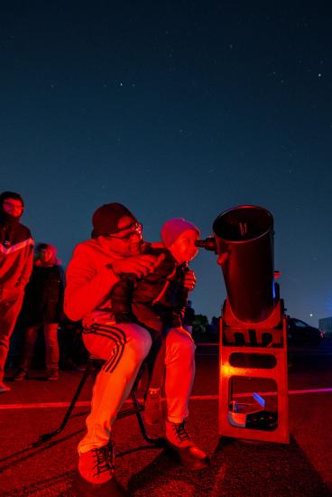 An child looks through a telescope with a thrilled expression while sitting on an adult's lap.