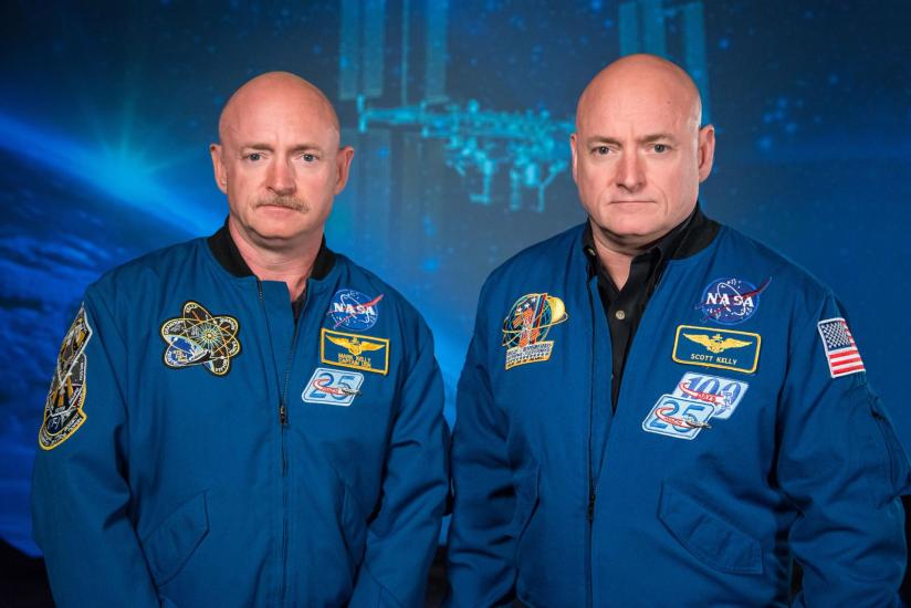 Two adult men who are identical twins stare at the camera. They are both bald, the one on the left has a mustache. They wear blue flight jackets with NASA patches on them. 