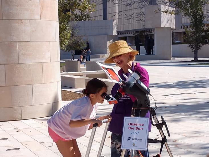 A person looking through a telescope outside during the day with a second person speaking to them.