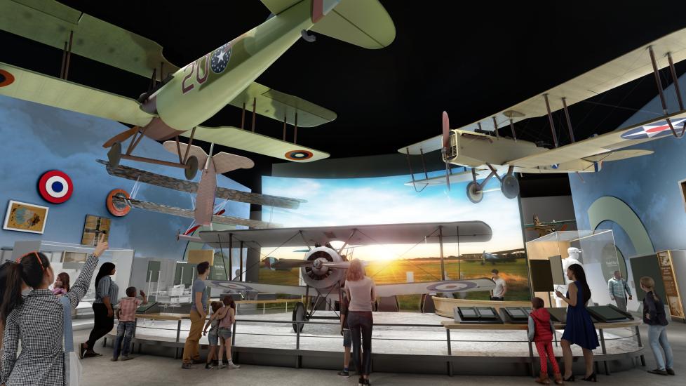 A digital rendering of a museum gallery shows several people standing in front of a display of three vintage biplanes. Behind the airplanes is a huge, floor-to-ceiling projection screen.