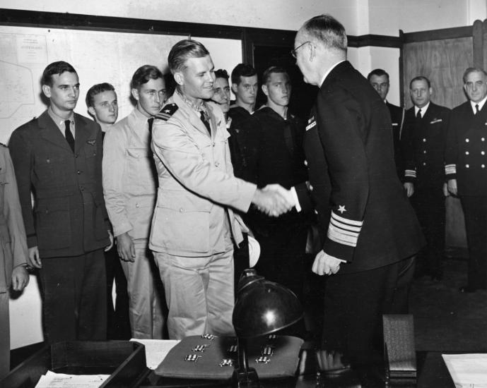 Two men in uniform shake in a room. A group of men in uniform stand behind them.