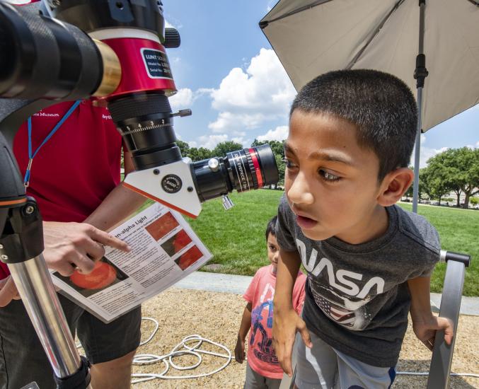 A young visitor in a NASA t-shirt looks through a telescope on a beautiful sunny day outside. 