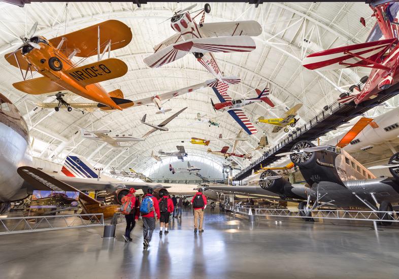 Planes both hand from the ceiling and stand on the ground in an aircraft hangar.