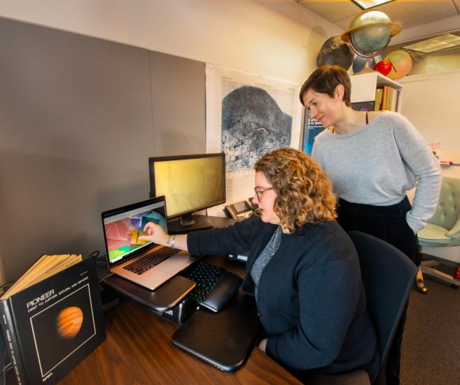 Two researches look at a computer monitor with colors displayed on it. In the background is a topographical map of a planet and globes of different planets. 