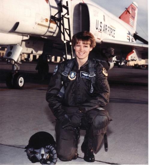 A woman posing next to an aircraft in a military flight suit.