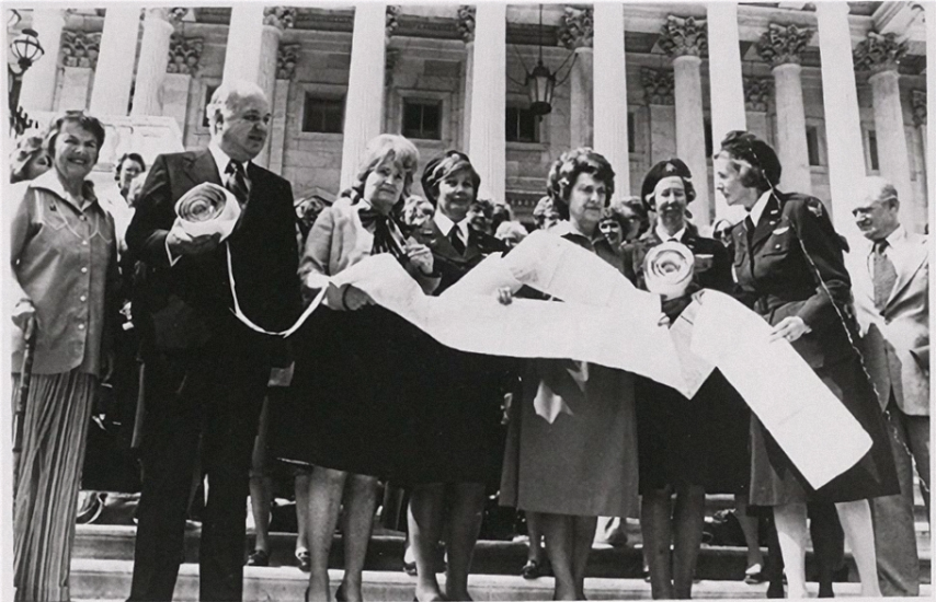 A group of men and woman holding up a long petition outdoors.