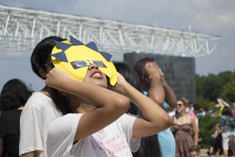 A young woman looks through a pair of eclipse glasses framed by a cutout of the sun.