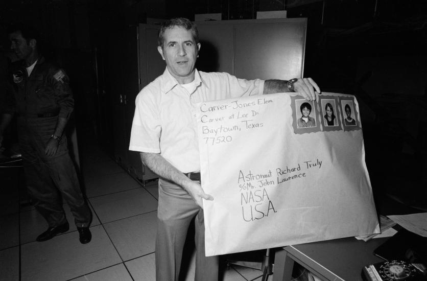 A middle aged man holds a giant envelope the size of an oven. The envelope is address to Richard Truly at NASA and is from Carver-Jones Elementary School. The "stamps" on the envelope feature three children in school portraits. 