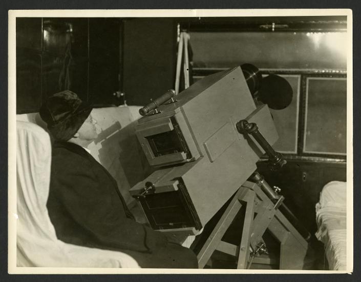 A person sits by the window of an airship with a large camera device pointed at the window.