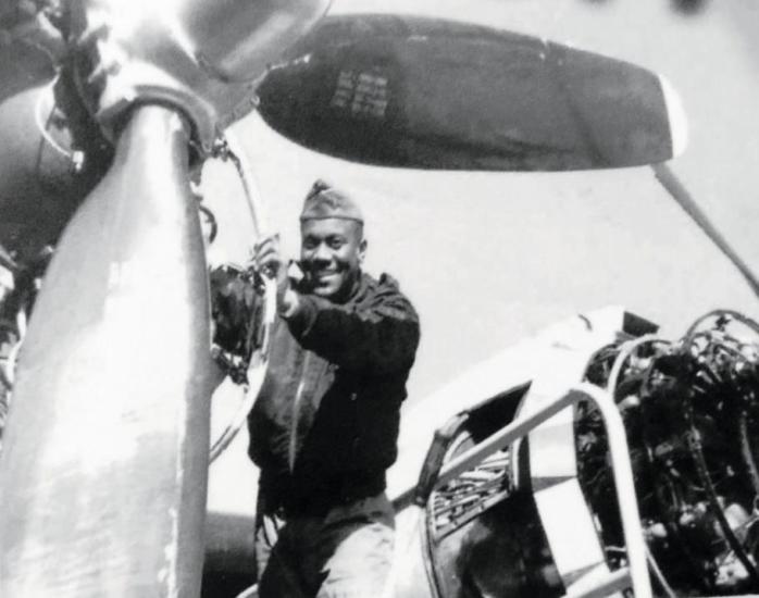 A man in a military uniform faces the camera and smiles as he works on an airplane propellor. 