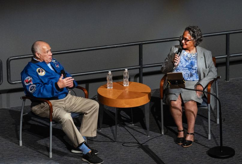 Two people in conversation on stage. On the left is Frederick Gregory, a former astronaut. He's a senior African American man and wear's a blue NASA flight jacket. He leans back in his chair, gesturing with his hands, mid-story. On the right is Dr. Cathleen Lewis, a museum curator. She's a middle aged African American woman in a grey suit. She holds a microphone and listens intently to Gregory. 