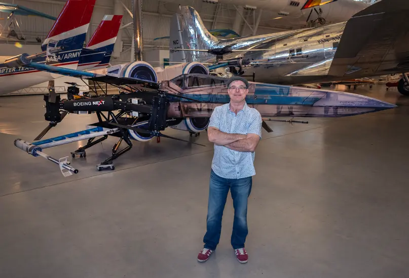 A middle-aged man in a cap, blue short sleeve button up, and jeans, stands with his arm crossed in front of a drone styled to look like a life-size x-wing from Star Wars.