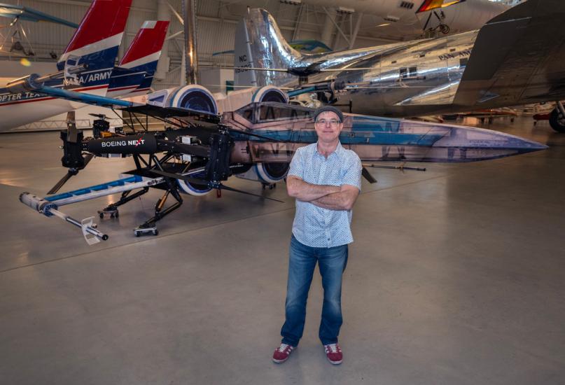 A middle-aged man in a cap, blue short sleeve button up, and jeans, stands with his arm crossed in front of a drone styled to look like a life-size x-wing from Star Wars.