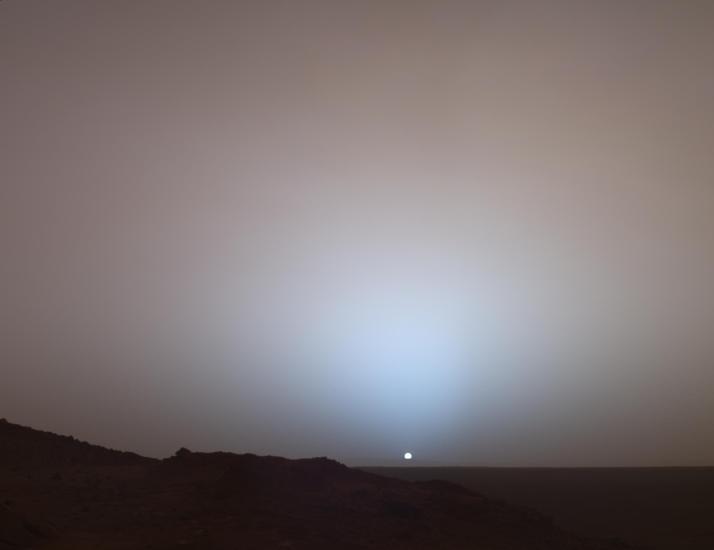 Mars. A landscape under a hazy sky with the sun visible as a small, bright dot near the horizon. The terrain appears to be barren and rocky. The atmosphere is diffusing the sunlight, creating a soft glow around the sun.