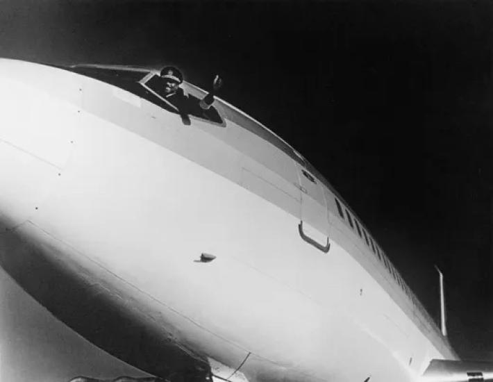 A photo of a man in a pilot's uniform leaning out of the window of an aircraft flight deck as he gives a thumbs up.