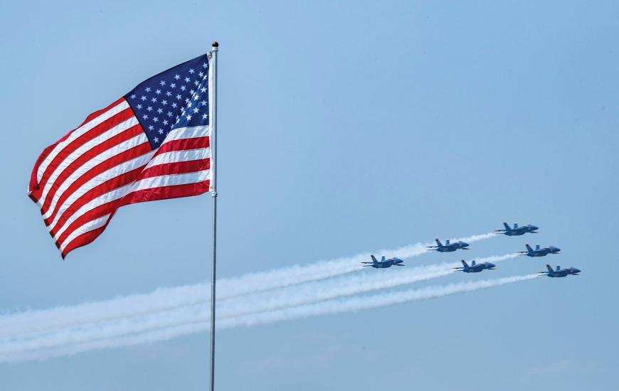 Six fighter jets in a trinagluar formation fly past an America flag attached to a long silver pole. White airshow smoke is streaming from the six jets during the afternoon flight.