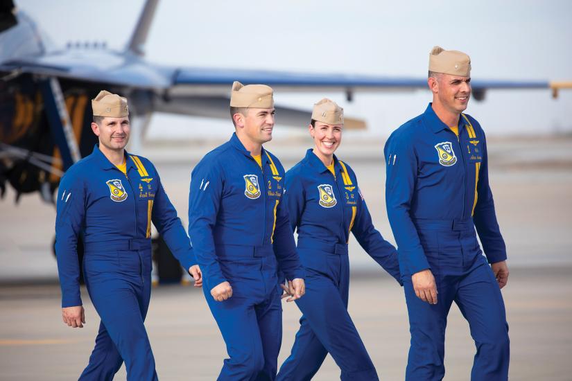 Four white people wearing blue flightsuits and military hats walk along the concrete ramp of a military air base. Three of the people are men; one of them is a woman. The woman is smiling.