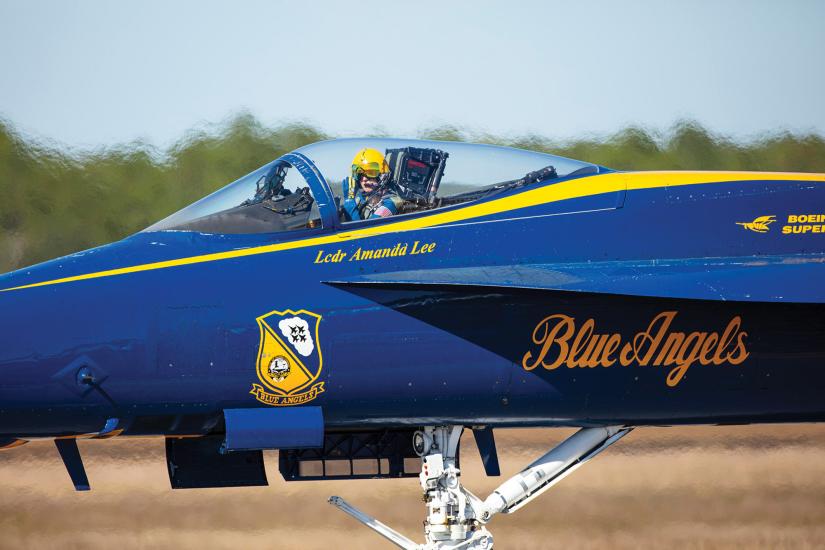 A person wearing a military helmet is sitting in the cockpit of a blue military jet. Painted on the side of the jet in yellow letters is "Blue Angels" and "LCDR Amanda Lee."