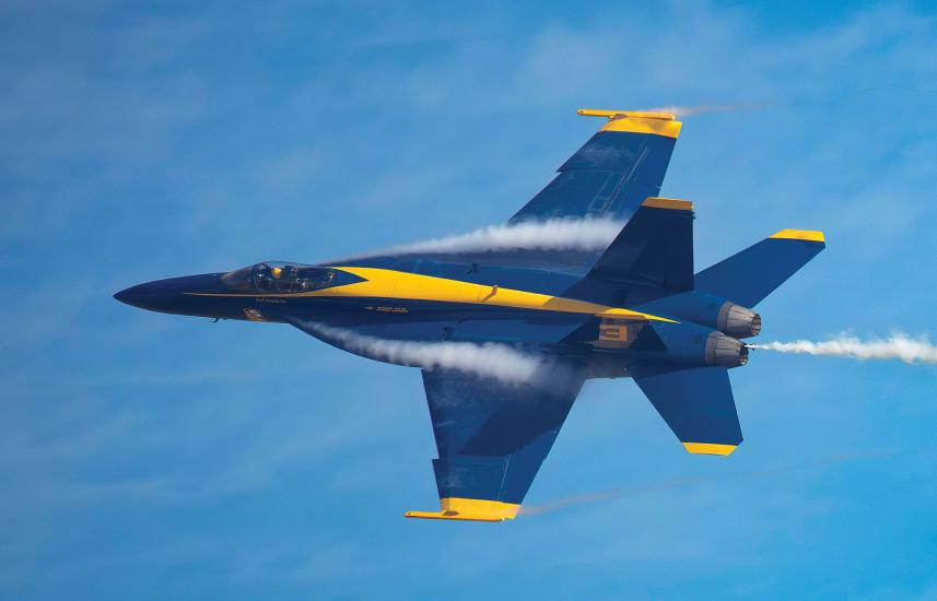 A blue military jet with yellow markings flies across a blue sky. The jet has twin engines, and the number 3 is painted on the side of the fuselage. Vapor streaks across the aircraft's wings, making its speed and percission evident. 