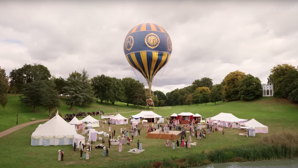 A vibrant hydrogen balloon with intricate blue and gold designs floats above a bustling outdoor event set against a cloudy sky. Below, several white tents are set up in a grassy park, with groups of people mingling and walking around.