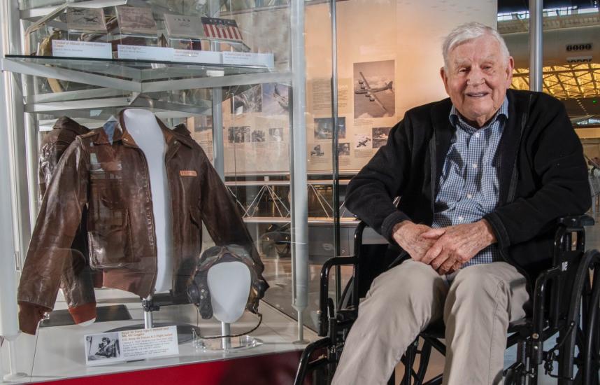 An elderly individual in a wheelchair smiles while seated next to a display of aviation memorabilia, including a leather jacket and a pilot helmet, inside a museum setting.