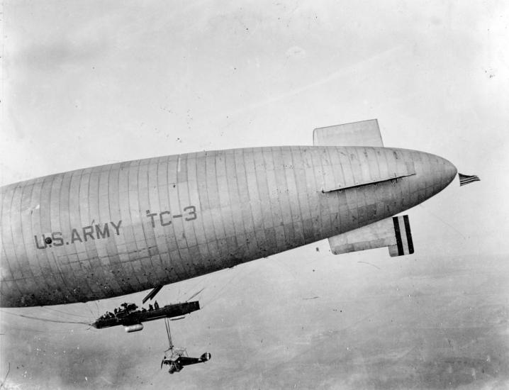 A large airship with US ARMY TC-3 written on the side a small aircraft hangs under the airship.