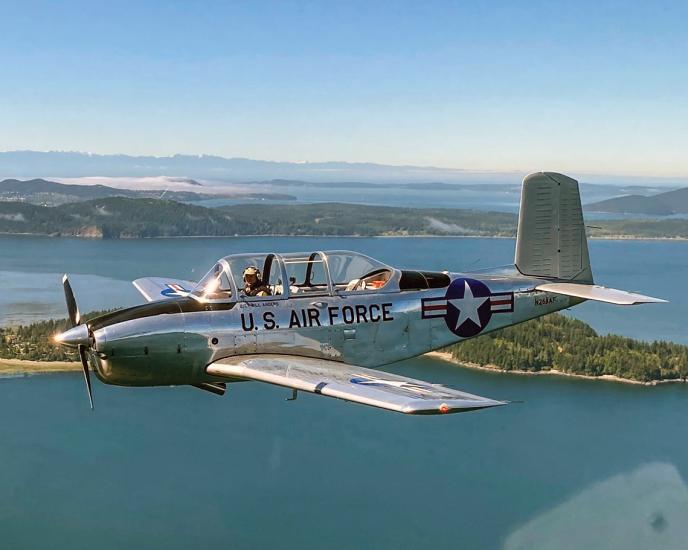 A small, single-engine airplane with a large glass canopy flies at low altitude over water and tree-covered islands. Mountains are in the background. On the side of the polished aluminum fuselage are the words "U.S. Air Force" along with a stars-and-bars USAF marking.